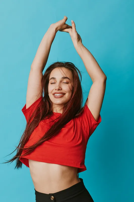 Young woman with long hair wearing red crop top and black pants posing with raised arms and closed eyes against blue background. Female model smiling and enjoying moment with carefree expression.