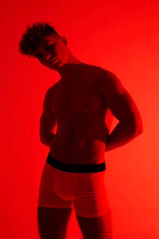 Male model, shirtless and muscular posing under red lighting with hands behind back, wearing white underwear against solid background for fashion or fitness photo shoot.