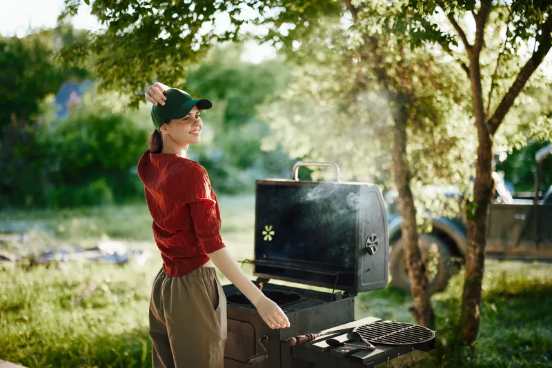 Cheerful woman grilling outdoors on a sunny day, wearing a green cap and casual clothes, enjoying summer cooking with a smoker and lush greenery in the background