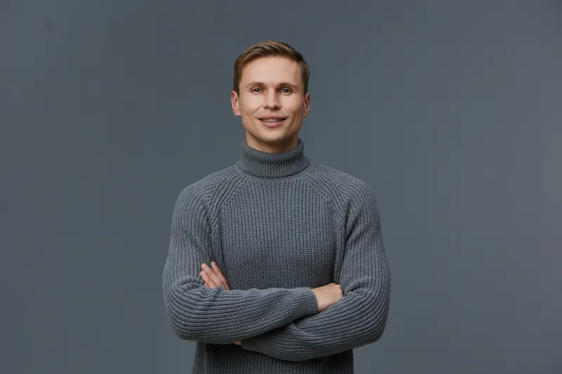 Smiling young man with blond hair wearing gray turtleneck sweater smiling confidently on gray background. Studio portrait of cheerful male model with cross arms, casual clothing, and friendly expressi