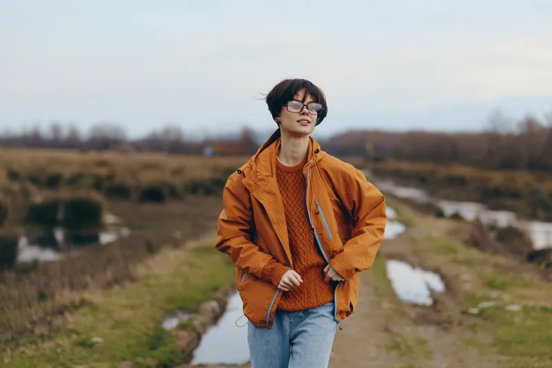 Woman lifestyle outdoors on trail in spring countryside, orange jacket and jeans, sunglasses, relaxed travel pose, nature fields and river valley, scenic hills in soft golden light, wide sky