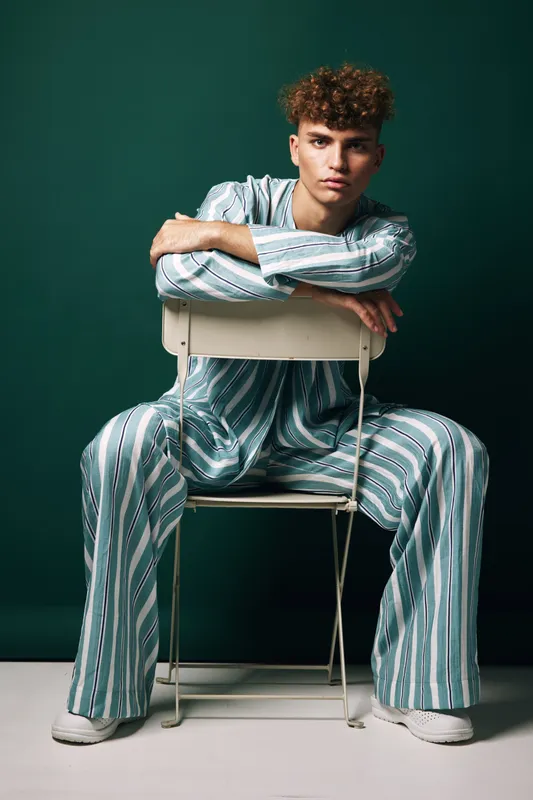 Young man with curly hair wearing striped pajamas sitting on chair backwards against green background. Casual fashion, relaxed pose and confident expression in studio setting.