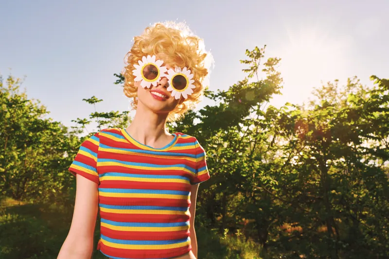 Cheerful woman outdoors, wearing flower sunglasses and striped shirt, sunlit moment, playful pose, vivid colors, lush greenery background, retro vibe, carefree mood, natural light