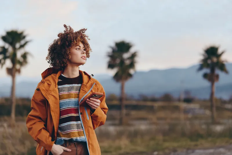 Woman lifestyle outdoors portrait with curly hair wearing orange jacket and striped sweater, holding phone, standing near palm trees, warm sunset light, calm thoughtful mood