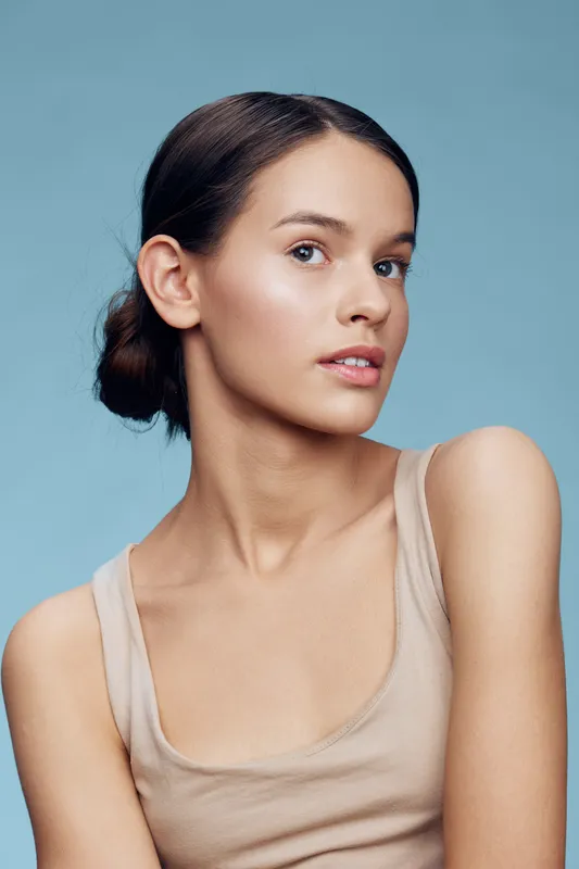 Young woman with a natural look, wearing a beige top, soft expression, against a calming blue background, showcasing beauty and confidence