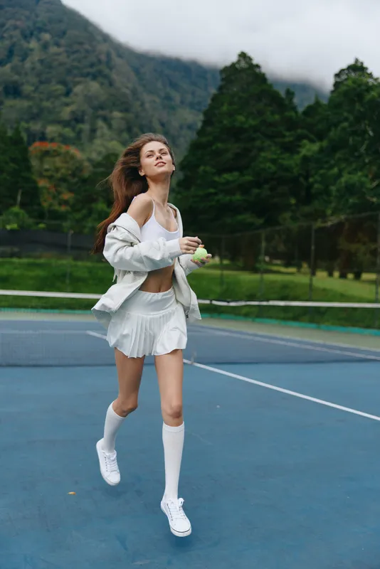Athletic young woman in white sportswear joyfully running on a tennis court, showcasing freedom and energy with a lush green background, perfect for fitness and healthy lifestyle concepts