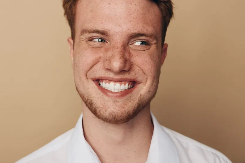 Smiling young man with freckles and red hair looks to the side. Close-up portrait on beige background. Concept of happiness, youth, and natural beauty with casual style.