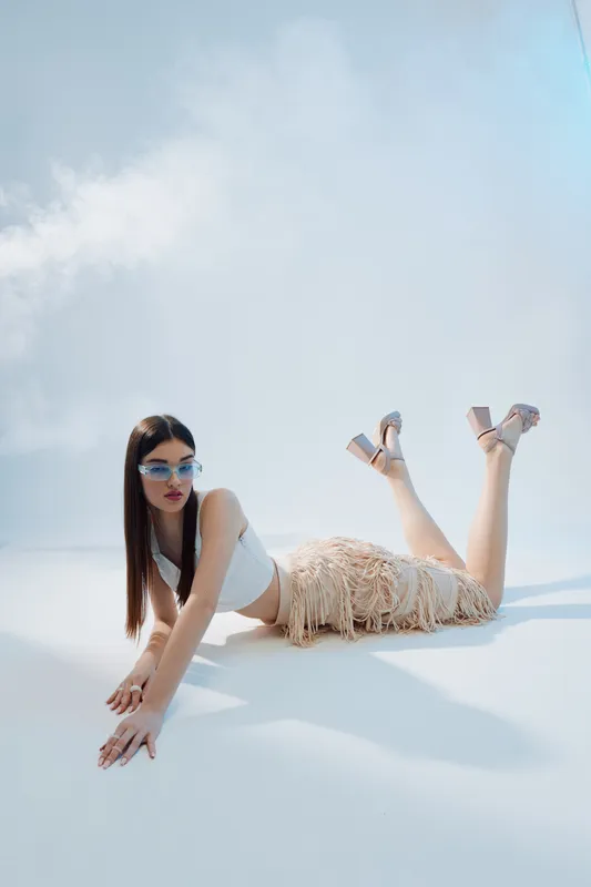 Young woman in stylish fashion sunglasses and a fringed skirt poses lying on the floor in bright studio lighting. Concept of beauty, fashion, and modern style with a minimalist background.