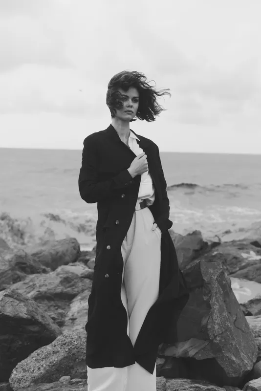 Monochrome fashion scene with a wind swept woman in a long coat and wide trousers standing on rocky shores by the sea, bold contrast and moody atmosphere.
