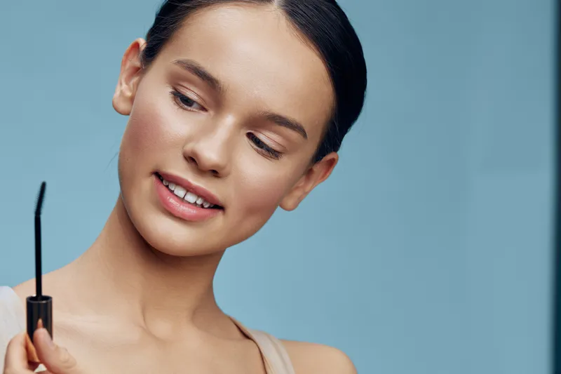 Cheerful young woman applying mascara, showcasing natural beauty and elegance in a soft blue background for beauty and makeup concepts