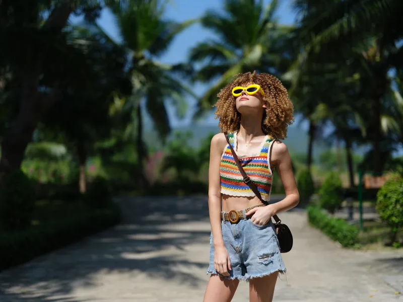 Smiling woman in yellow sunglasses enjoying sunny tropical day wearing colorful top and denim shorts with palm trees in background