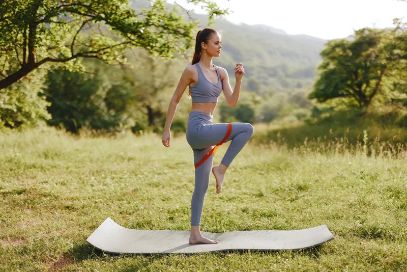 Young woman exercising in nature wearing gray sportswear on a yoga mat, showcasing a healthy lifestyle and fitness concept in a vibrant outdoor setting
