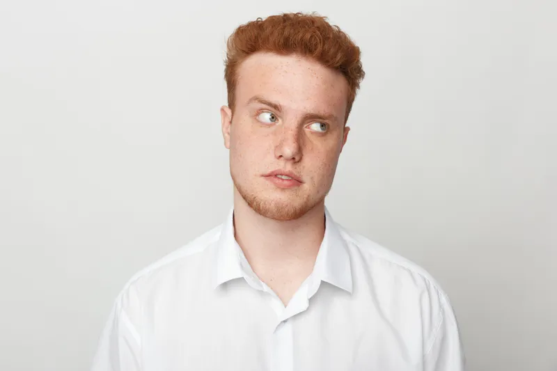 Redhead young man in white shirt looks sideways with thoughtful expression. Studio portrait on light background. Concept of contemplation, decision making, and casual style.