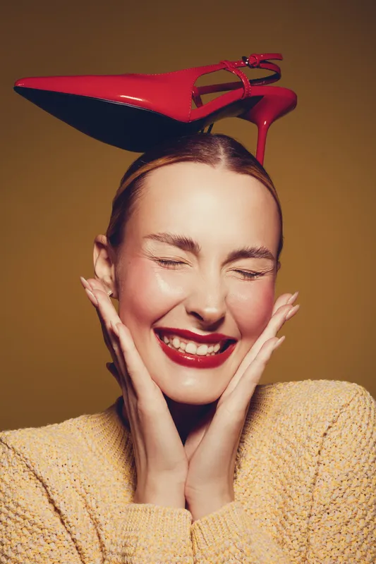 Smiling woman with closed eyes holds face with hands while balancing red high heel shoe on head. Studio portrait with warm lighting and yellow background. Concept of fashion and joy.