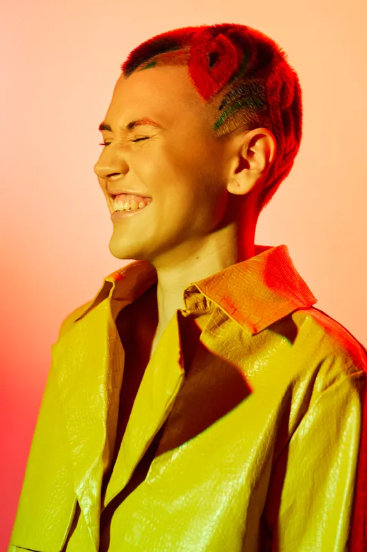 Smiling woman with colorful braided hair and yellow jacket enjoys vibrant warm lighting, expressing happiness and style in a modern fashion portrait.