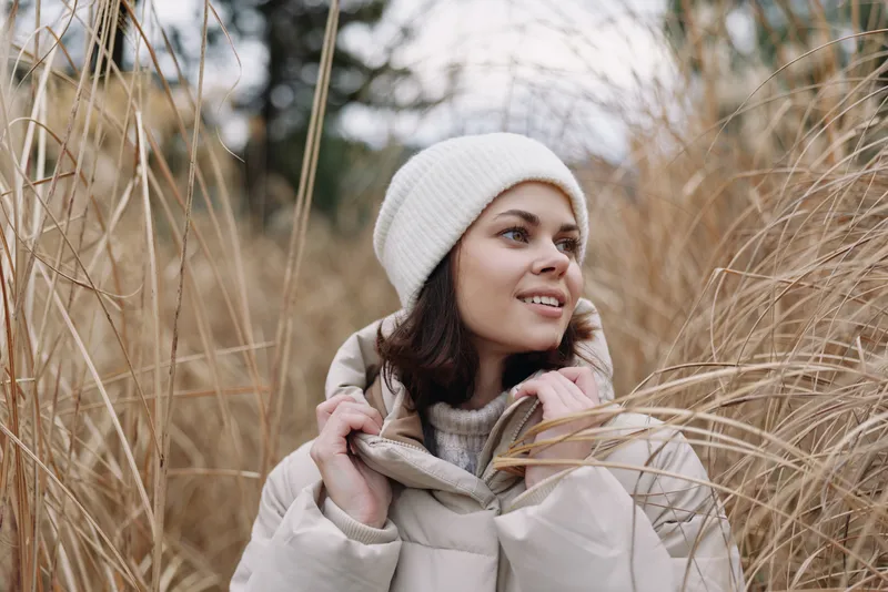 Woman wearing a warm coat and white beanie stands among tall grasses in a chilly outdoor setting, creating a serene winter portrait with natural textures, soft light, and calm mood.