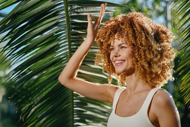 Smiling woman with curly hair enjoying tropical sunlight near green palm leaves, wearing white tank top, expressing happiness and natural beauty outdoors.