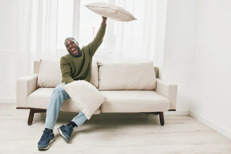 Young Black man joyfully playing with cushions in a bright living room, showcasing a sense of happiness and relaxation