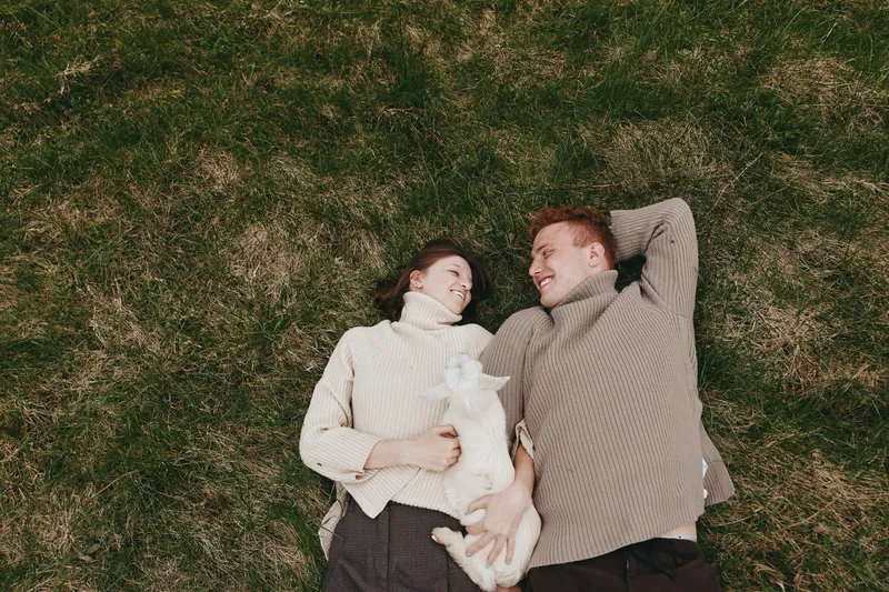 Couple and cat resting on grass outdoors in a relaxed lifestyle scene, smiling faces, warm light, cozy sweaters, intimate moment, natural setting, playful mood, affectionate connection, candid