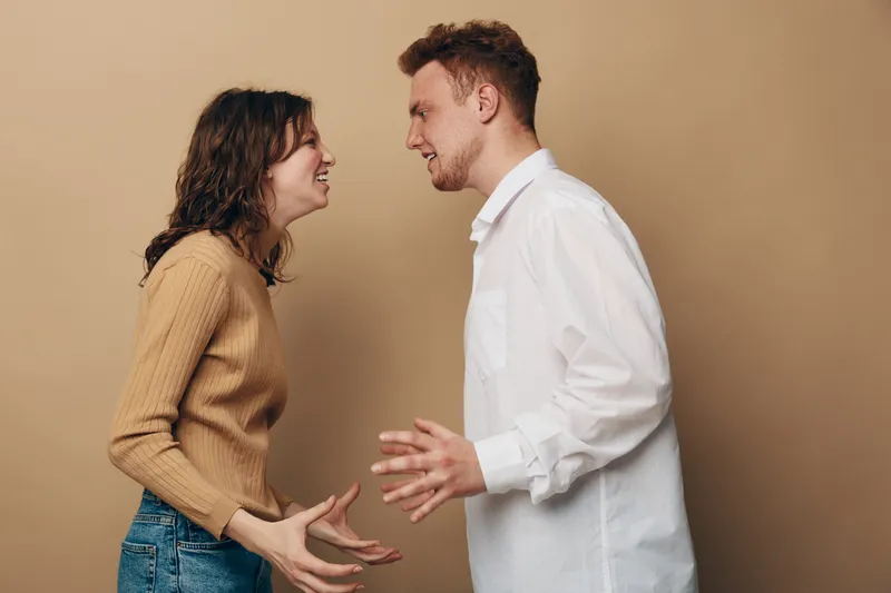 Young man and woman argue facing each other with intense expressions and gestures. Casual clothing and neutral background emphasize conflict and emotional tension between couple.