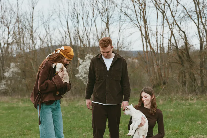 An outdoor family moment with a young goat kid in a field, one adult kneels to pet the kid while others smile nearby, creating a candid lifestyle scene of animal companionship and rural bonding.
