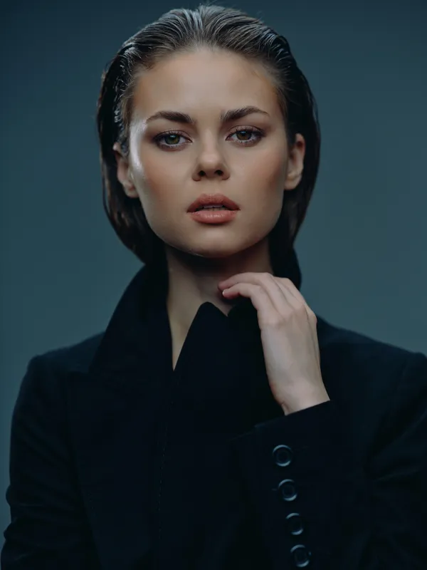Close-up portrait of a confident woman with slicked-back hair wearing a black jacket and a serious expression on a dark background.