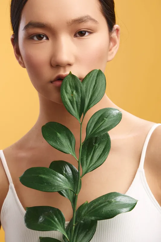 Asian woman holds a vivid leafy plant close to her face in a studio portrait, conveying natural beauty, serene wellness, and subtle elegance through soft lighting, gentle shadows, and a clean