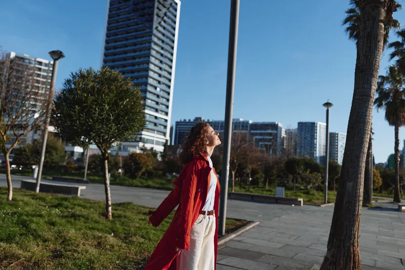 A stylish woman in a red coat walks through a sunny urban park, with palm trees and tall buildings rising behind her, conveying confidence and contemporary city life.