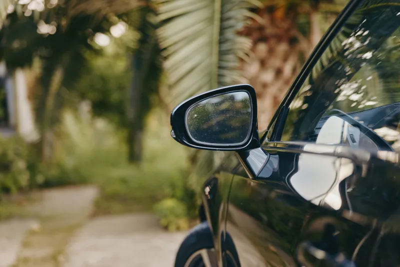Car side mirror on black car with clear reflection, parked on a driveway near palm trees, closeup of exterior detail and glossy paint under natural light