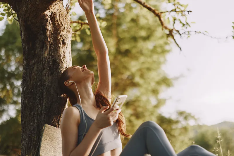 A young woman enjoying a sunny day outdoors, dressed in activewear, reaching towards a branch while relaxing in nature, showcasing a joyful and carefree mood