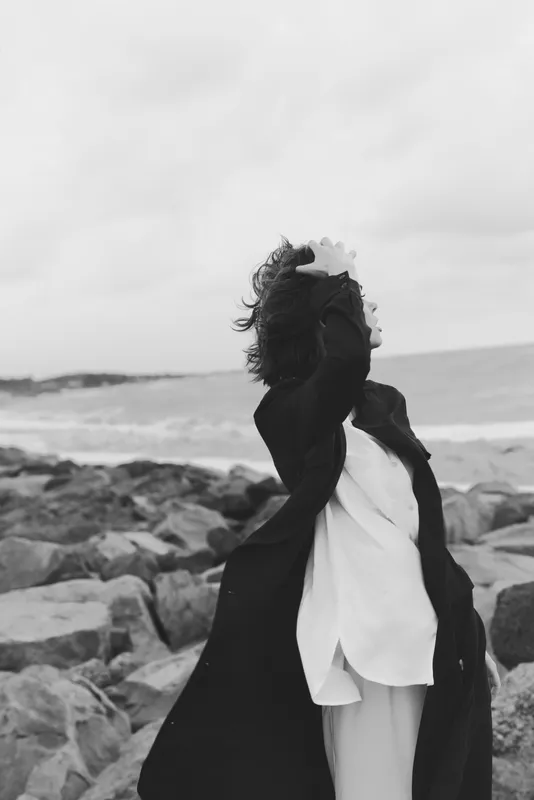 Coat dancing in wind by rugged shoreline, monochrome scene capturing movement and drama, outdoor fashion on rocky coast with sea spray and moody atmosphere, dramatic clouds overhead