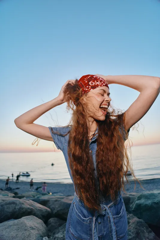 young woman laughing with long hair and red bandana on rocky beach at sunset with calm sea background and clear sky