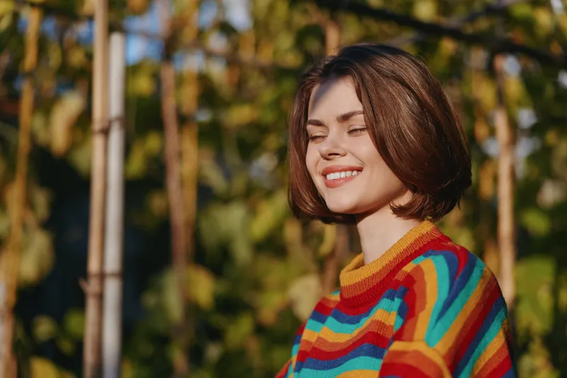 Woman smiling with eyes closed, young woman in her 20s wearing a colorful striped sweater, short hair portrait in sunny outdoor garden, casual lifestyle fashion with relaxed warm sunlight mood.