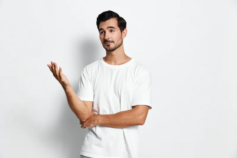 Casual young man in a white shirt gestures thoughtfully with a slight smile, standing against a solid pale background. Confident lifestyle concept.