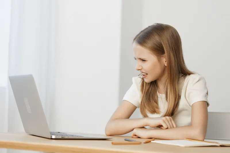 A frustrated young woman with long hair in a white blouse, looking at her laptop, conveys emotions of confusion and stress in a modern office setting