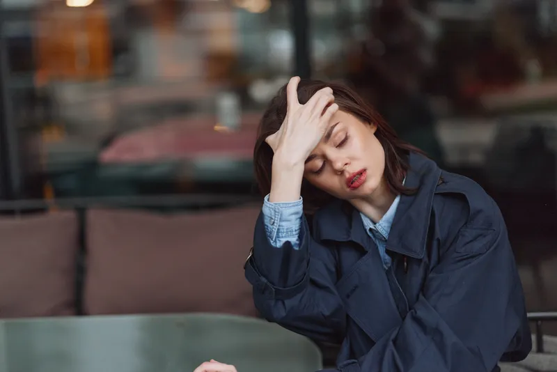 Tired woman with tousled hair rests her head on her hand at a cafe table, wearing a denim jacket. Casual portrait conveying fatigue, stress, and introspection in soft, warm light.