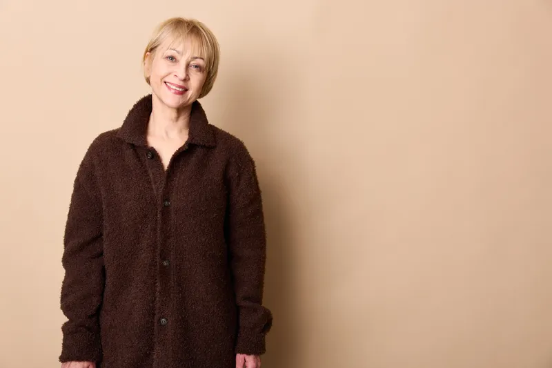 Elderly woman portrait, smiling, standing in dark coat, on beige background, studio indoor headshot, warm soft light, relaxed senior female, kind calm expression