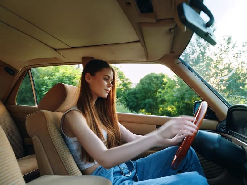 Young woman driving vintage car, enjoying a sunny day, dressed casually in a tank top and jeans, with a focus on freedom and adventure.