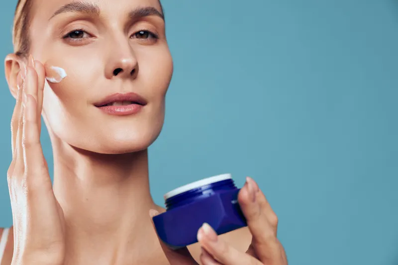 Woman applying face cream with fingers and holding a blue moisturizer jar Close up on blue background Skincare, self care, wellness, skin care, studio lighting