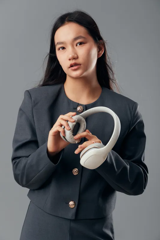 Young woman in gray business suit holding white headphones, posing against neutral background. Professional style with long black hair in studio, fashion concept with electronic accessory in hand.