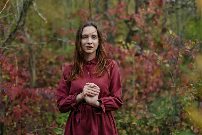 A young woman in a burgundy dress stands among autumn trees, facing the camera with a genuine, calm expression. An authentic natural portrait capturing honest emotion, texture and presence.