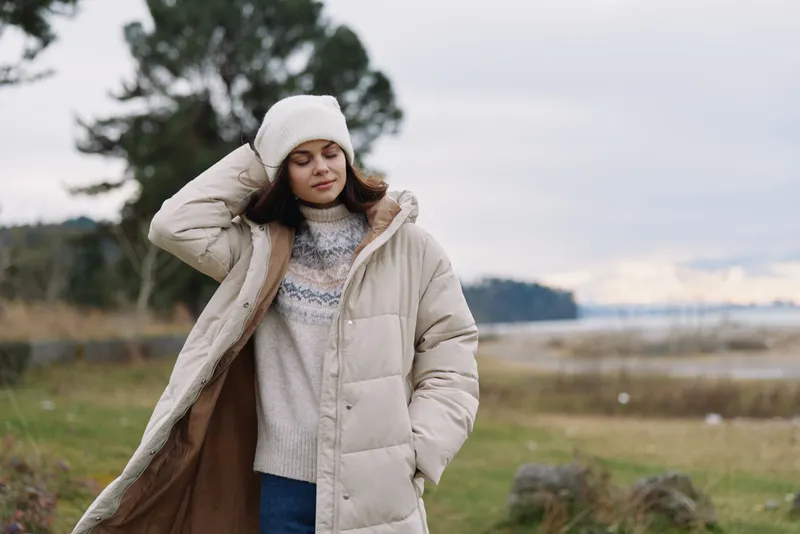 Woman in a beige puffer coat and knit sweater outdoors, wind ruffles hair, beanie hat, calm landscape with water and trees, chilly mood, candid fashion moment in nature