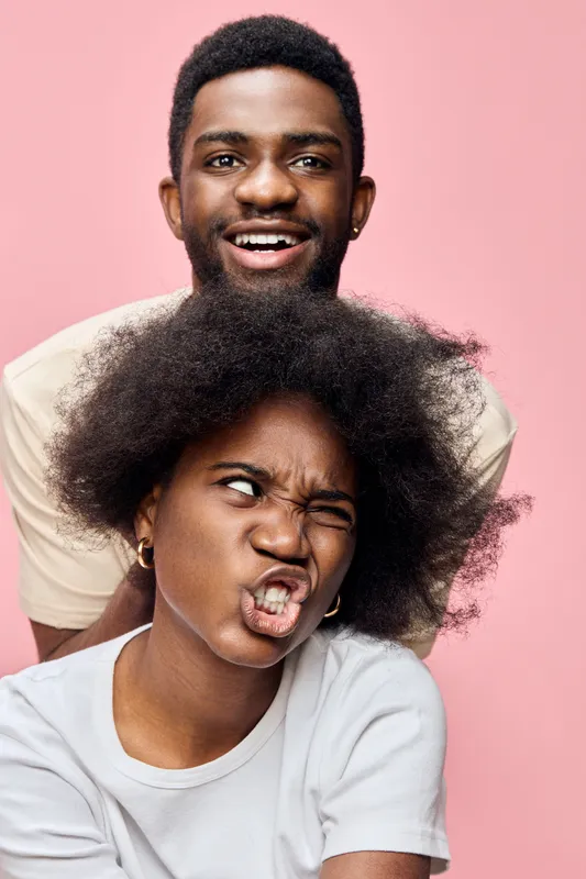 Joyful young black couple with playful expressions on a pink background, showcasing authenticity and spontaneous moments in a vibrant, youthful style