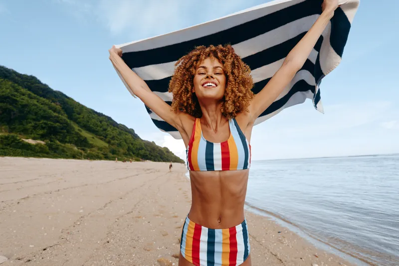 Young woman with curly hair on a sunny beach lifts a striped towel over her head, wearing a colorful striped bikini, smiling as the calm sea meets the sand.