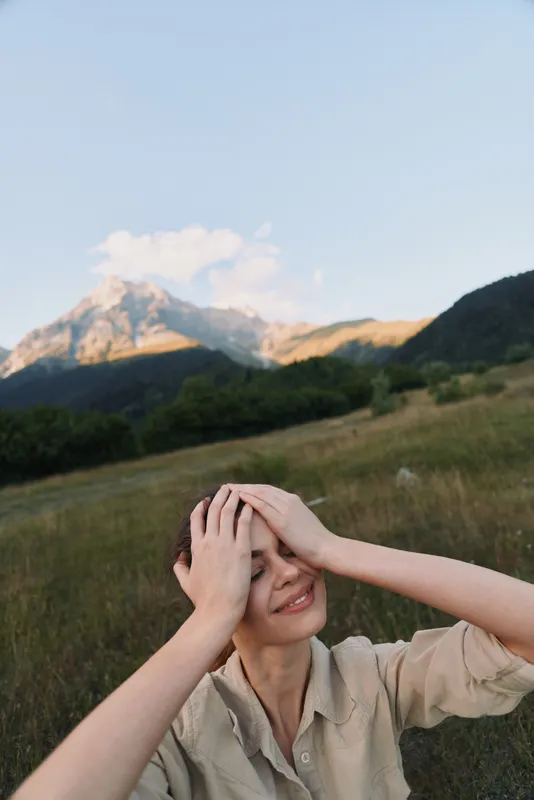 Cheerful woman outdoors, enjoying sunlight, relaxed pose, hands on head, smiling expression, natural colors, wide field scenery, distant mountains, calm breeze