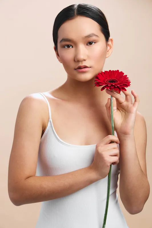asian woman holds a red flower near her face, wearing a light tank top, posed against a neutral background, exuding calm elegance and soft beauty in a studio portrait.