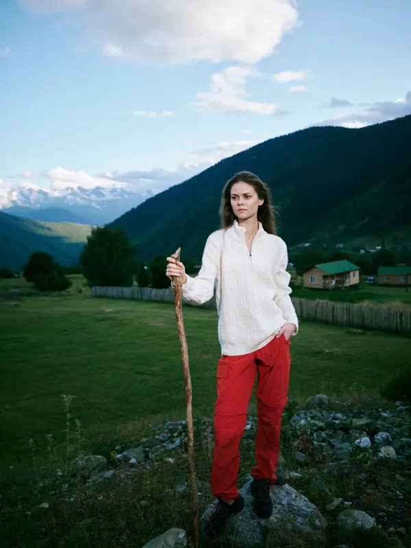 Outdoor scene of a woman standing on a rocky overlook with a wooden staff, mountains in the background, green valley, blue sky, casual white blouse and red pants, exploration and nature