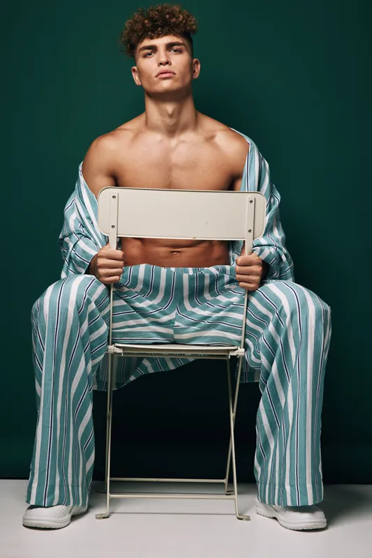 Young man with curly hair sitting on chair backward wearing striped pajamas with open shirt showing bare chest against dark green background in studio.