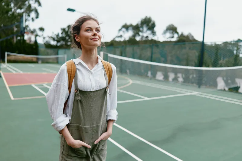 Young female tennis player in casual overalls, joyful expression on a tennis court, greenery in the background, showcasing a sporty, vibrant lifestyle and spring vibe
