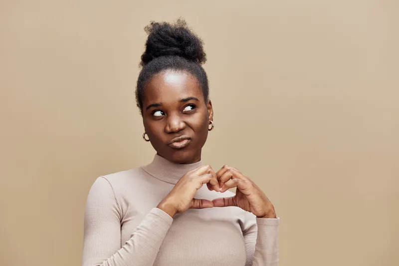 Young Black woman making a heart shape with her hands, expressing playful emotions against a beige background, perfect for love and relationship themes